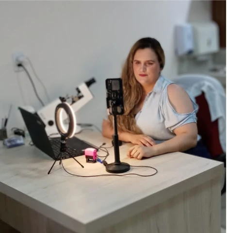 Dra. Gabriela à mesa do consultório, diante do notebook e do celular com anel de luz